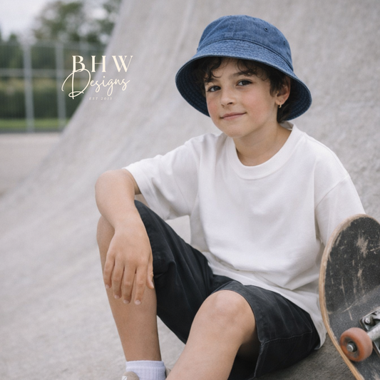 Child sitting on a skateboard wearing a blue bucket hat and white t-shirt at a skate park.