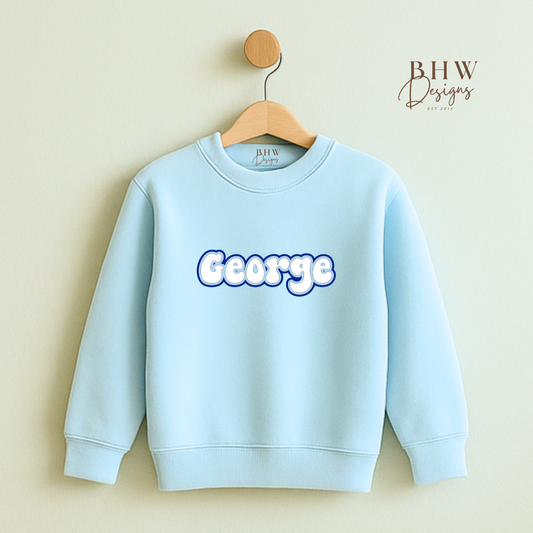 Light blue sweatshirt with 'George' printed on it, hanging on a wooden hanger against a beige wall.