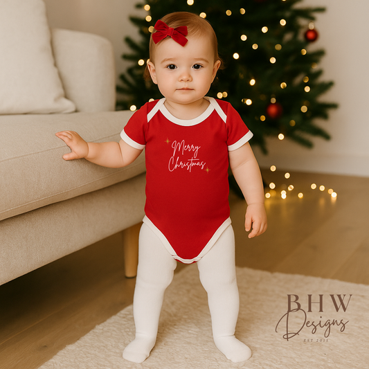 Baby wearing a red 'Merry Christmas' onesie in a home setting with a Christmas tree.