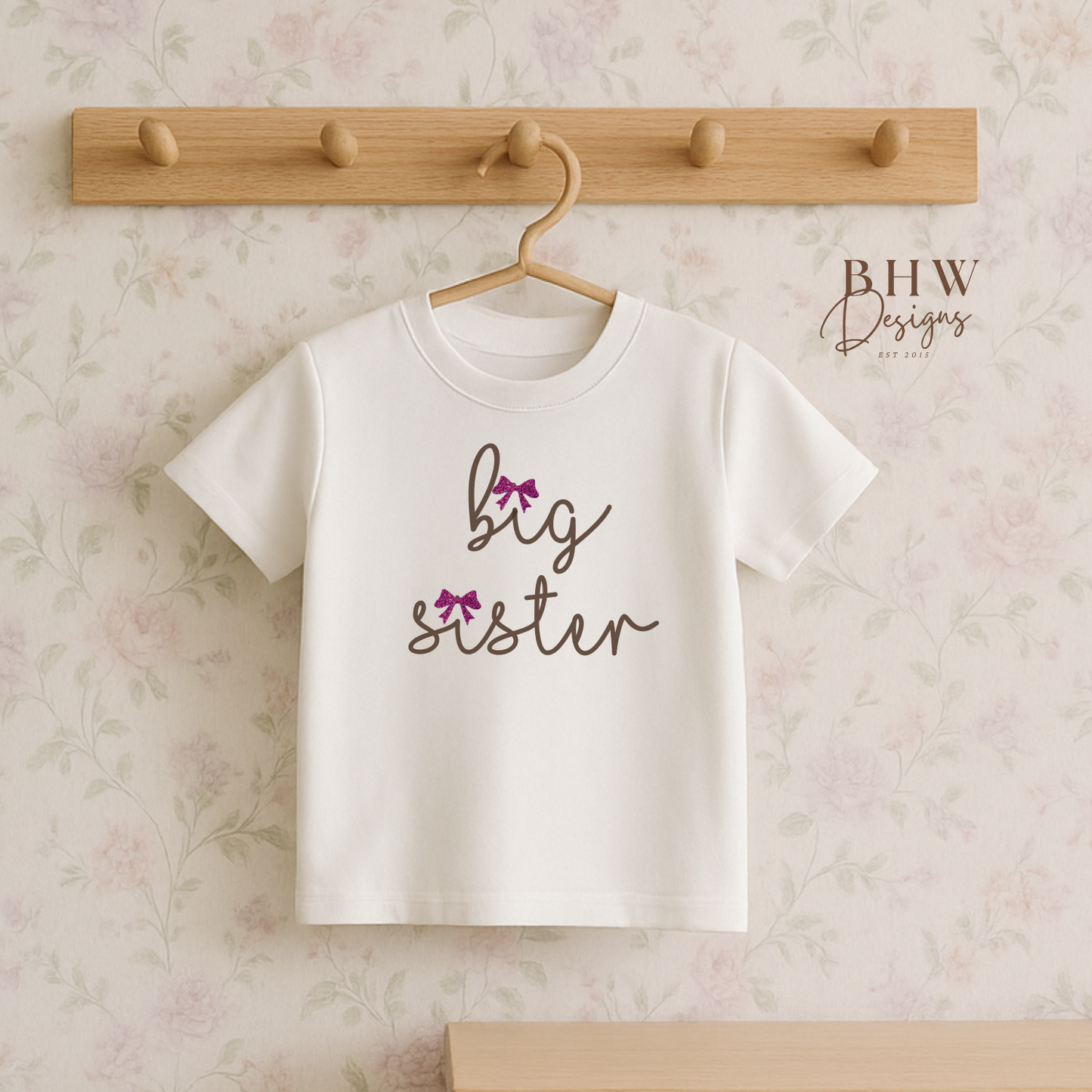 White t-shirt with 'big sister' text on a wooden hanger against a floral wall.