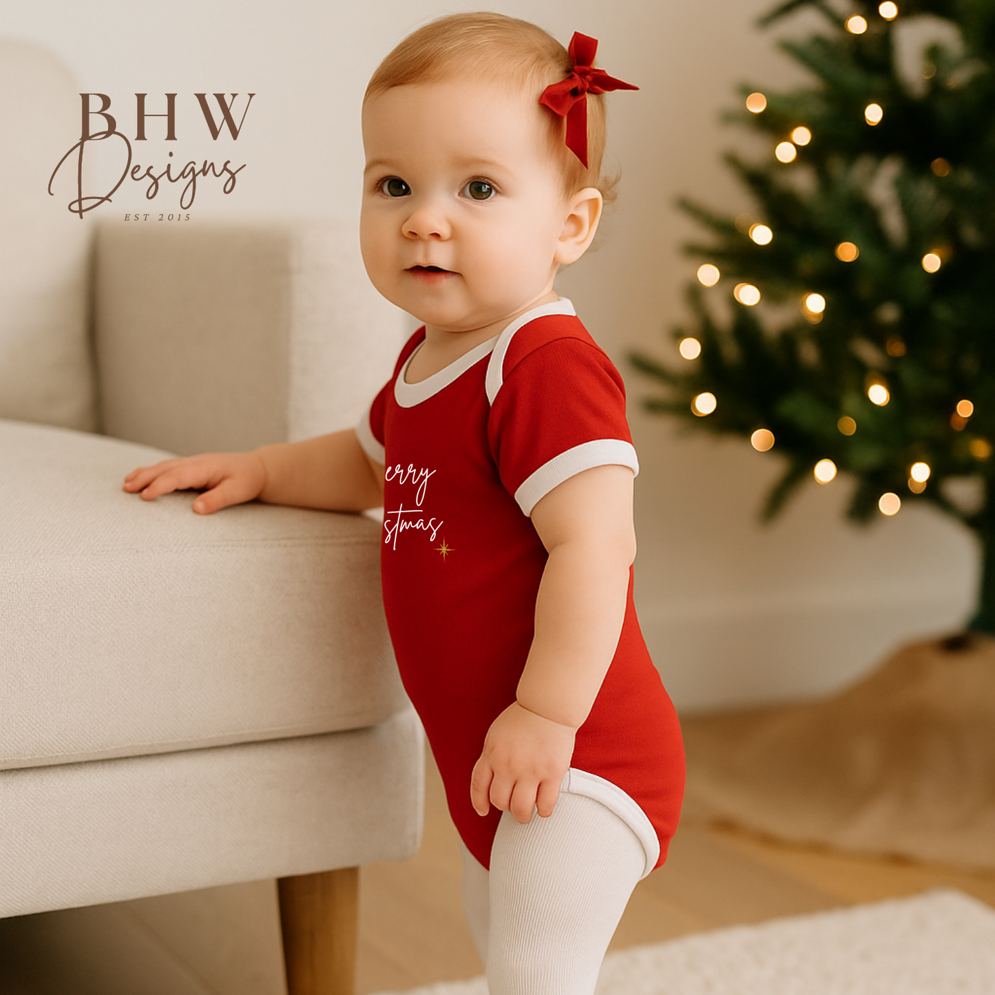 Baby in a red onesie with white tights standing in front of a decorated Christmas tree.