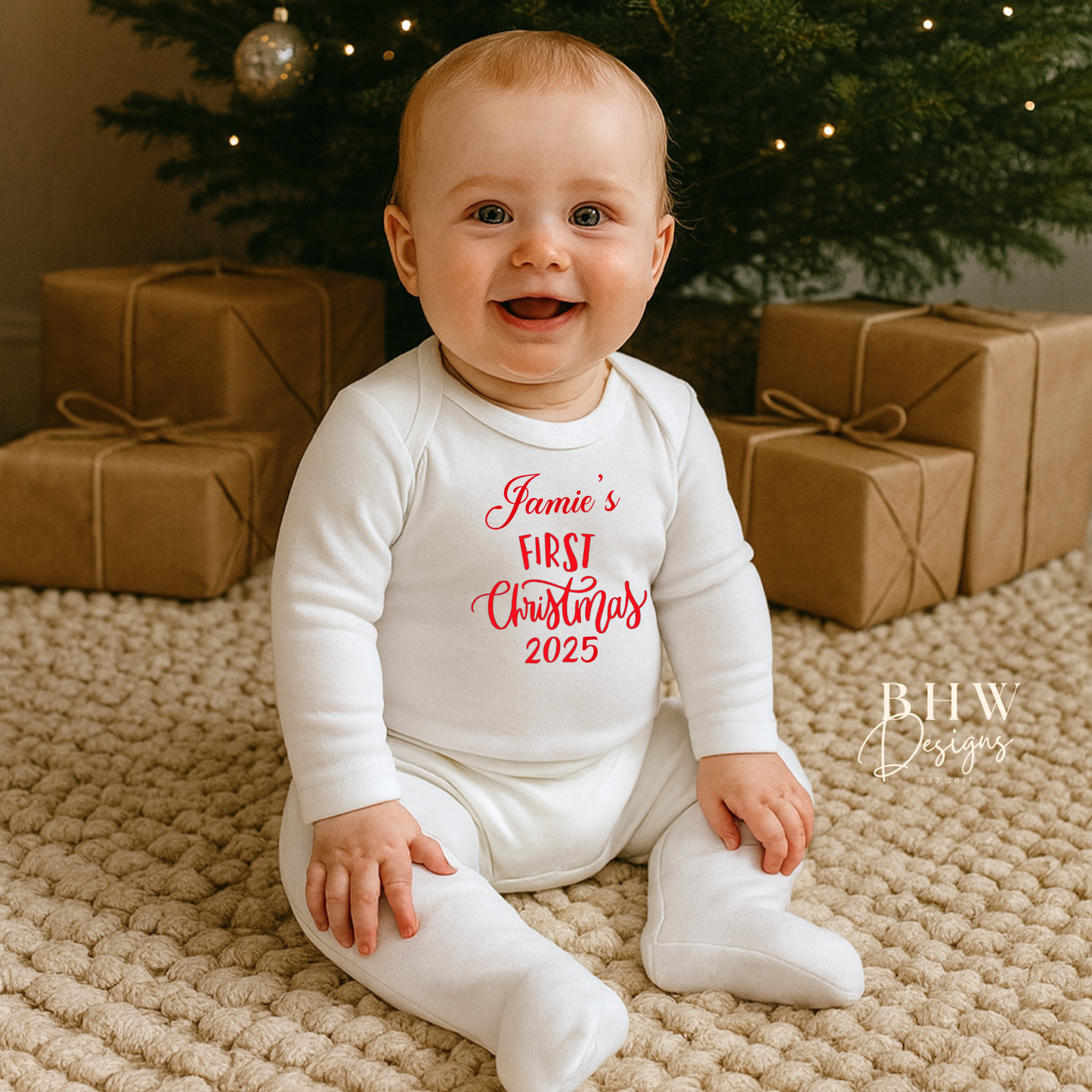 Baby in a 'Jamie's First Christmas 2025' onesie sitting in front of a decorated Christmas tree with presents.