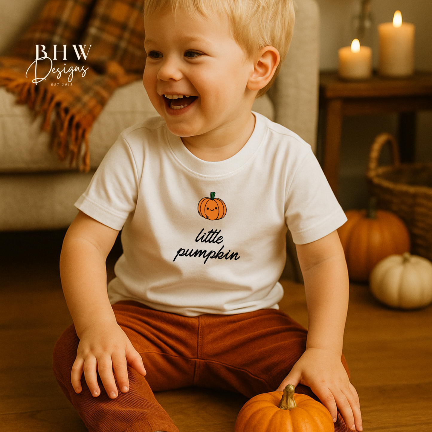 Child wearing a 'little pumpkin' shirt holding a small pumpkin in a cozy indoor setting.