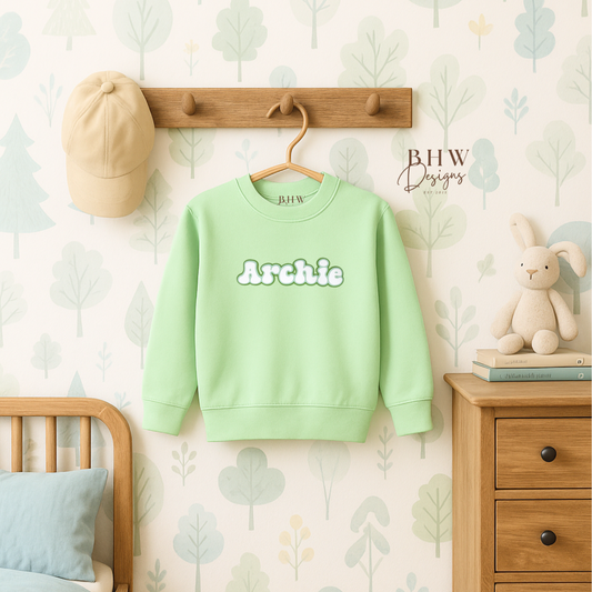 Light green sweatshirt with 'Archie' printed on it hanging on a hook in a child's room.