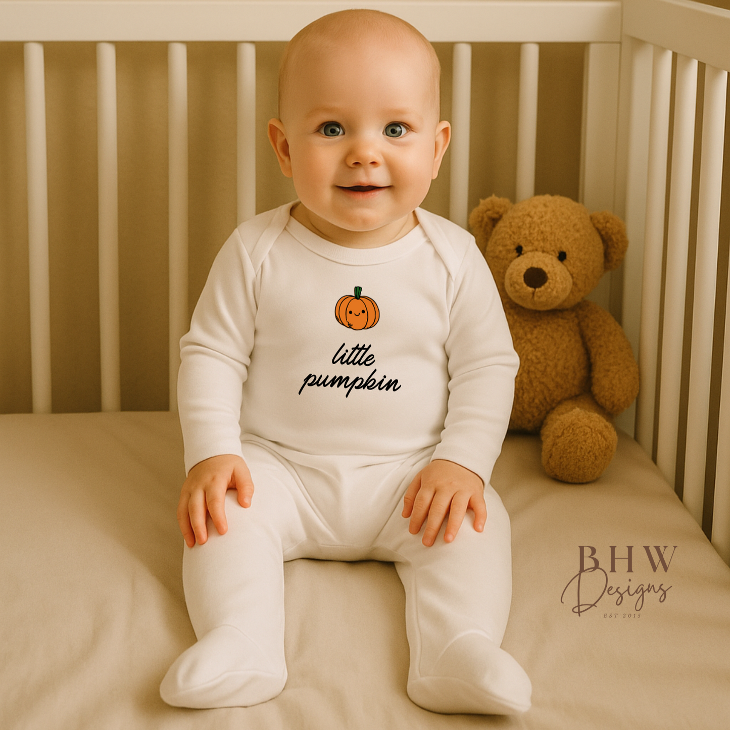 Baby sitting in a crib wearing a 'little pumpkin' onesie with a teddy bear in the background.