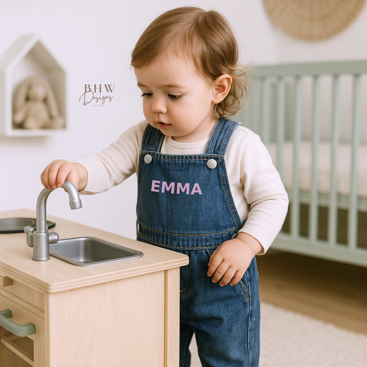 Child in denim overalls with 'EMMA' printed on them, playing with a toy kitchen sink.