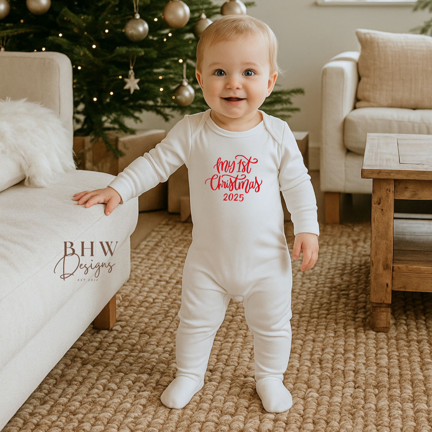 Baby in a white onesie with My 1st Christmas 2025 inred text standing in front of a decorated Christmas tree.