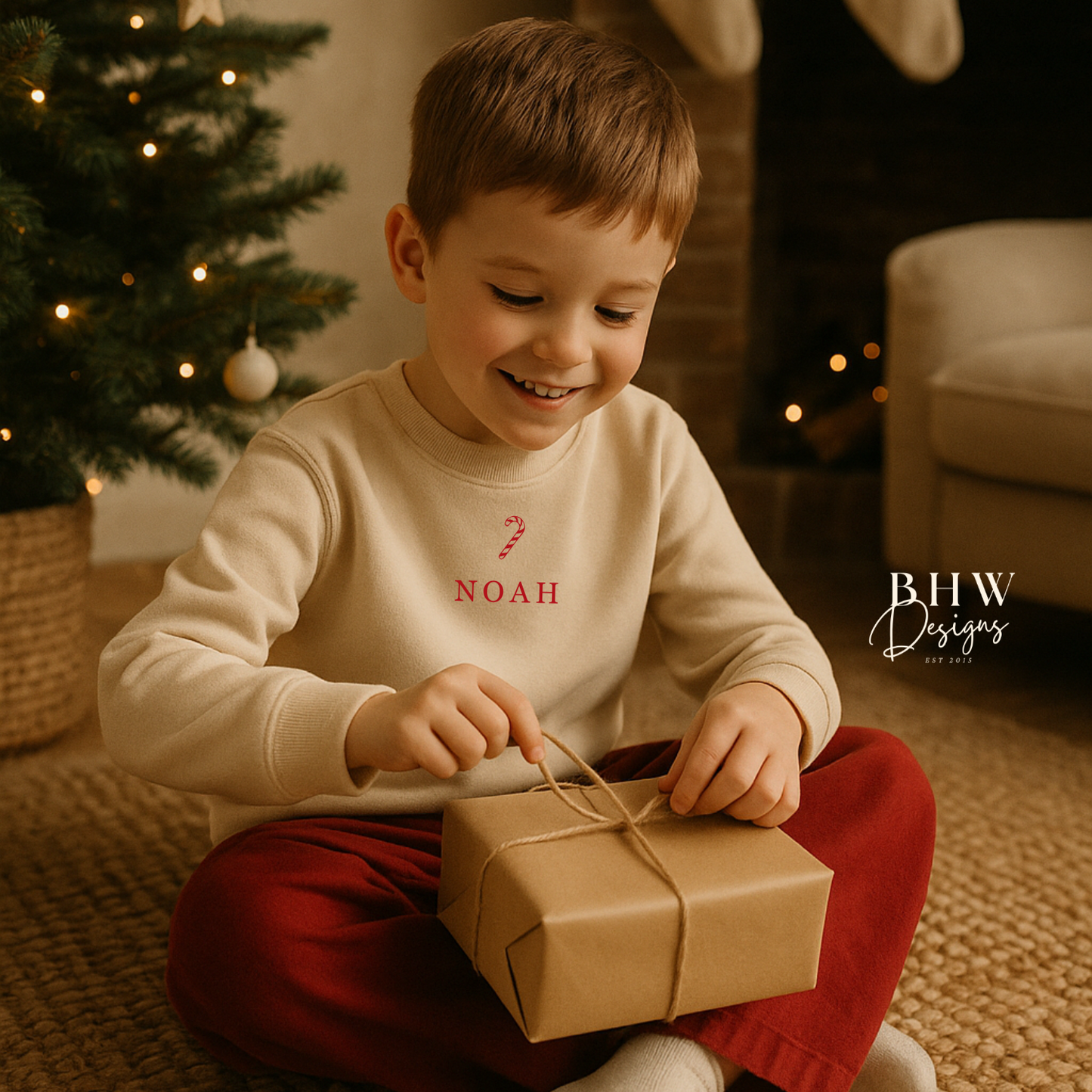 Child wearing an oat colour sweatshirt with a candy cane and Noah on the front in red opening a gift in a festive setting with a Christmas tree and stockings.