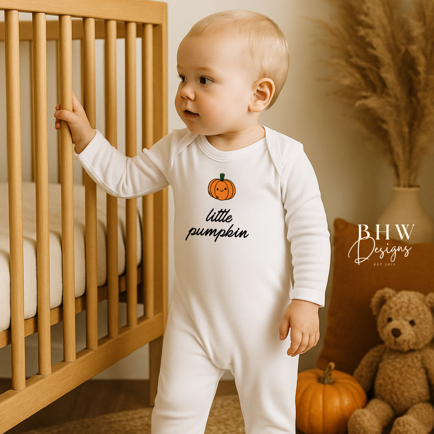 Baby in a 'little pumpkin' onesie standing next to a crib with pumpkins and a teddy bear.