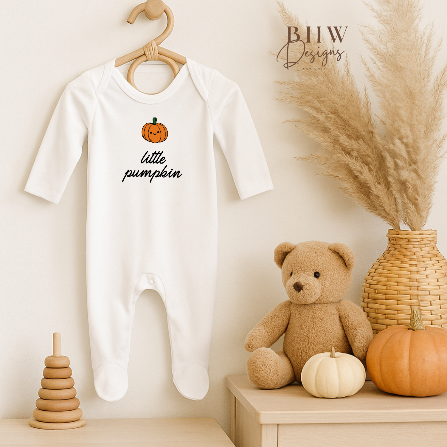 Baby onesie with 'little pumpkin' design on a hanger, surrounded by decorative items including a teddy bear and pumpkins.