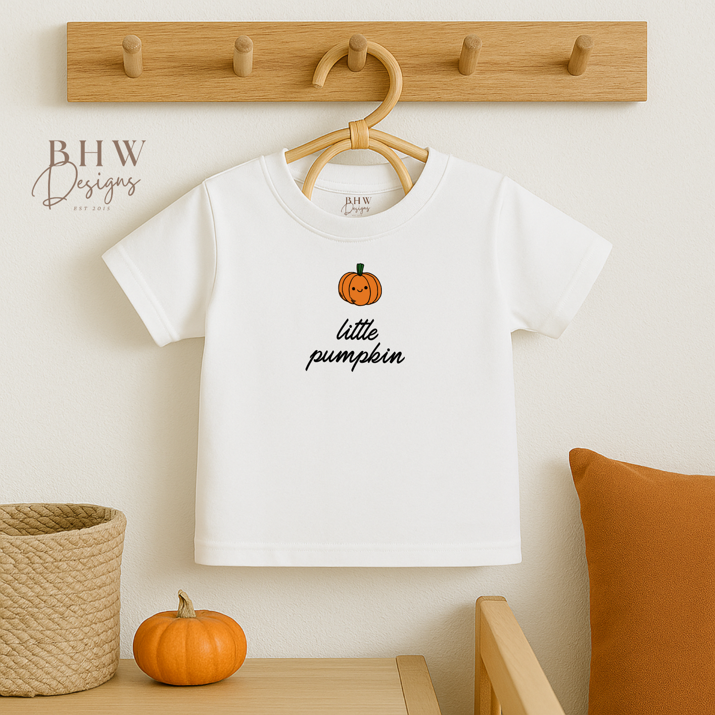 White t-shirt with 'little pumpkin' design on a hanger against a neutral wall.