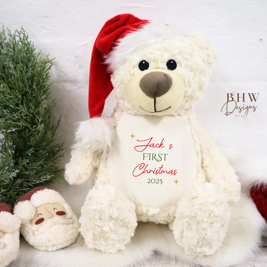 White teddy bear with a red Santa hat and 'Jack's First Christmas 2025' text, surrounded by Christmas decorations on a light wooden surface.