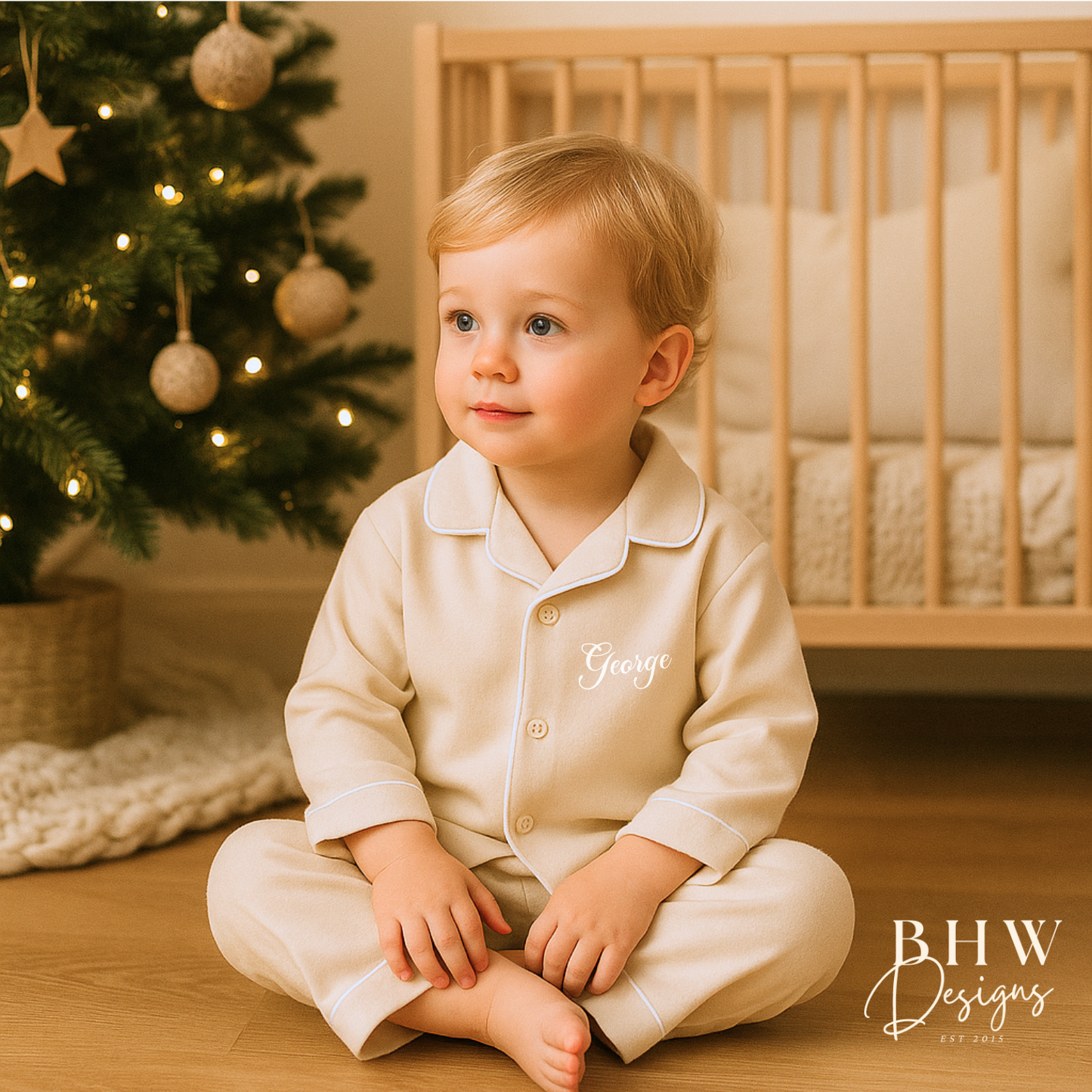 Boy in beige cotton pyjamas Personalised with George in white sitting in front of a decorated Christmas tree.