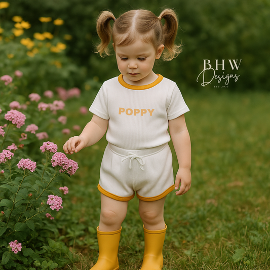 Child in a ribbed white outfit with 'Poppy' printed on it, standing in a garden with flowers and greenery.