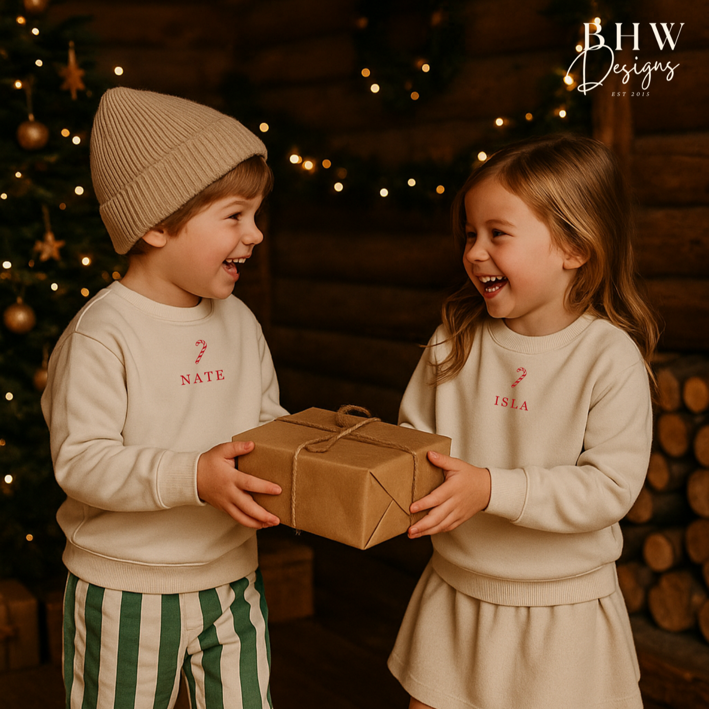 Two children exchanging a gift wearing oat colour sweatshirt with a candy cane and Nate and Isla on the front in red