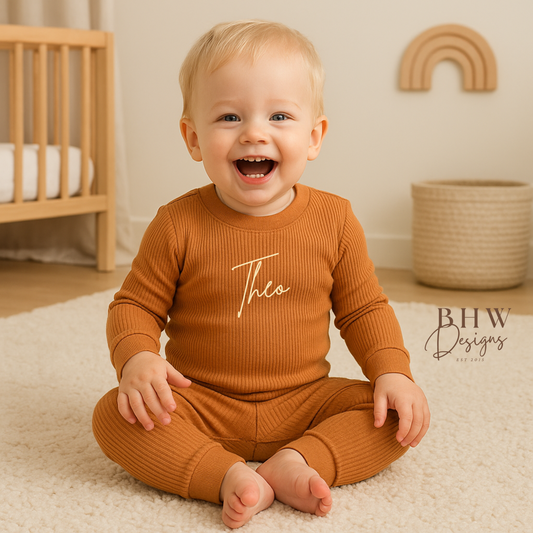 Baby wearing a rust outfit with 'Theo' printed on it, sitting on the floor in a nursery.