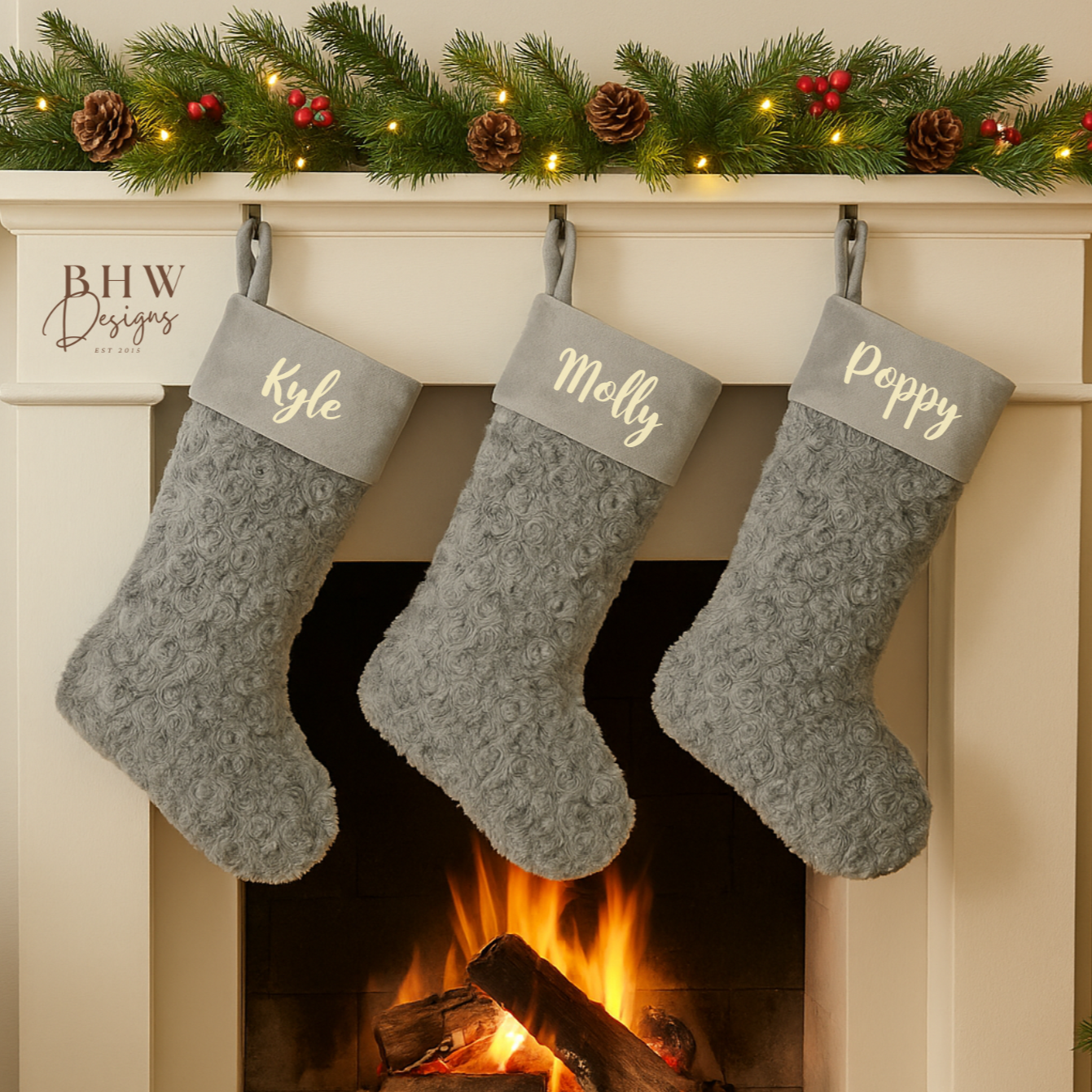 Three grey Christmas stockings labeled 'Kyle', 'Molly', and 'Poppy' hanging above a fireplace with a warm fire.
