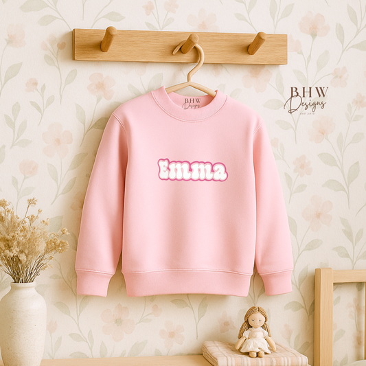 Pink sweatshirt with 'emma' text hanging on a wooden hook against a floral wallpapered wall.