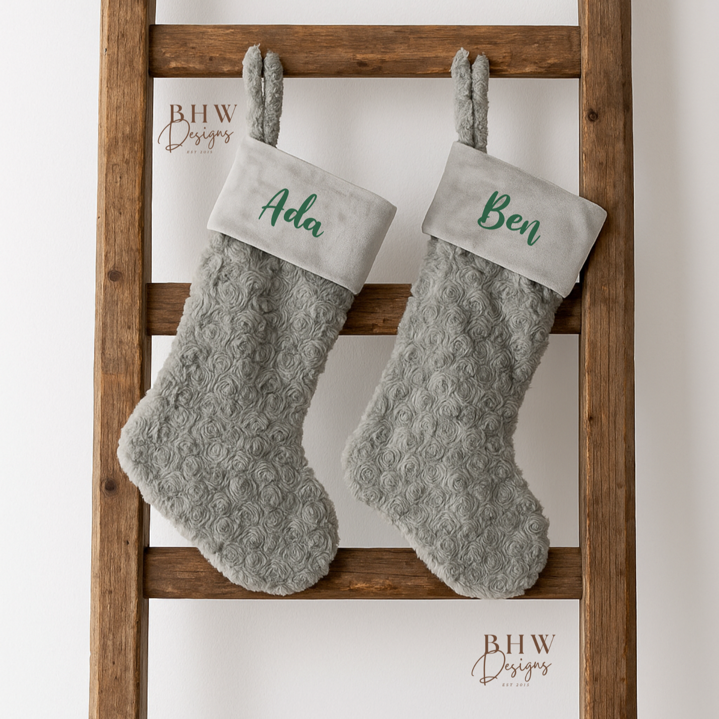 Two grey fluffy stockings with names 'Ada' and 'Ben' on a wooden ladder against a white background.