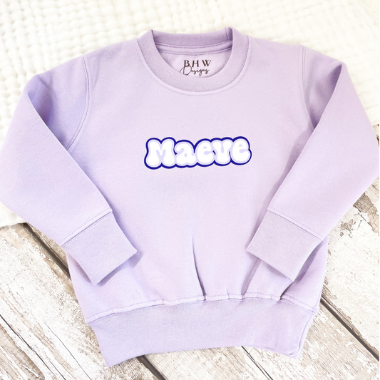A personalised long-sleeved lilac sweatshirt for children with the name Maeve printed in bold letters on the front.