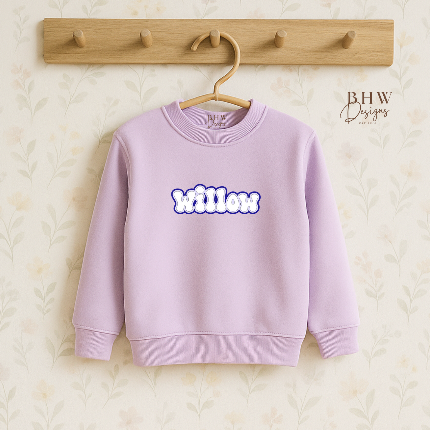 Lavender sweatshirt with 'Willow' printed on it, hanging on a wooden hook against a textured beige background.