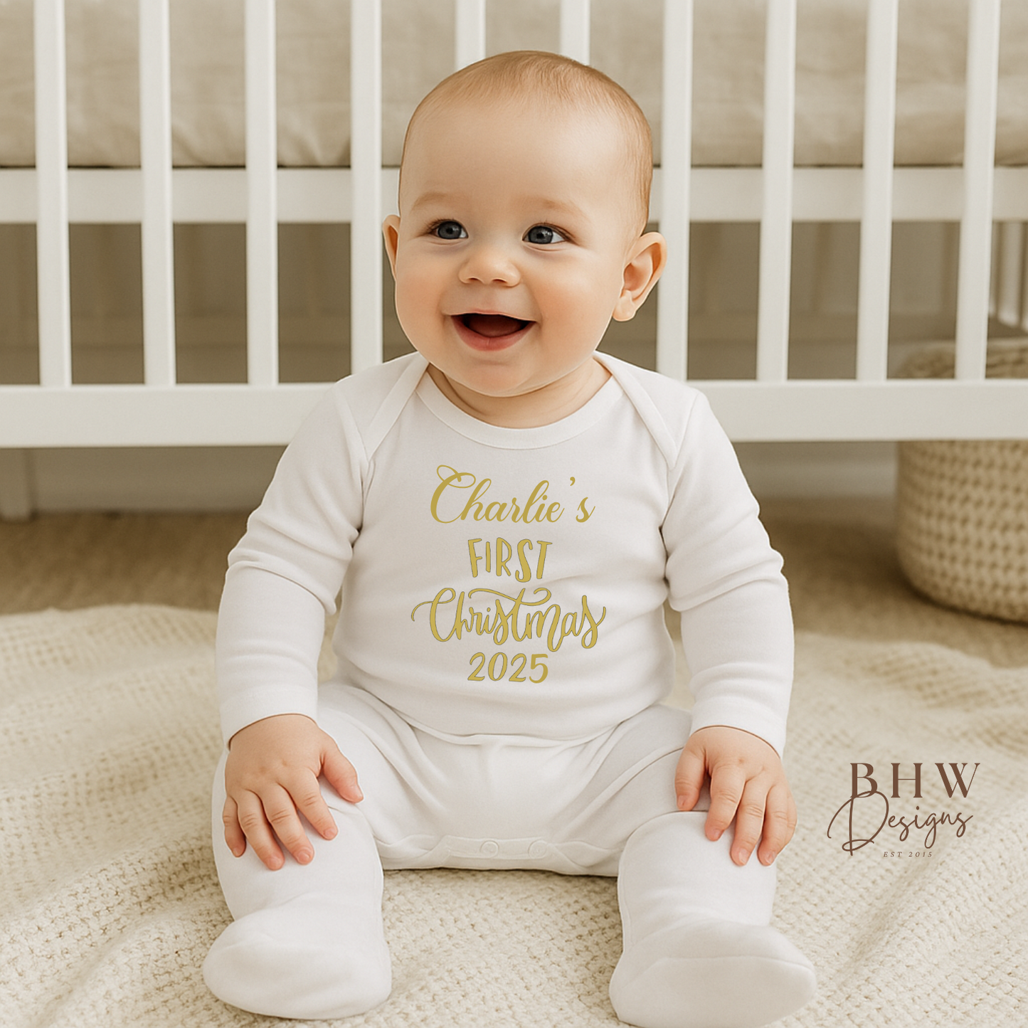 Baby wearing a white onesie with gold text saying Charlies first Christmas 2025, sitting on a blanket in front of a crib.