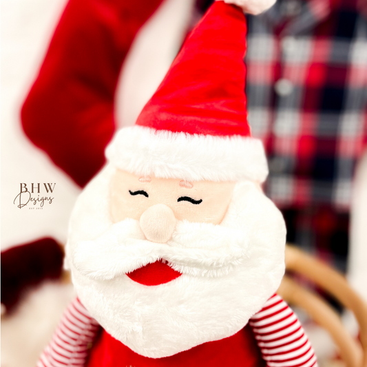 Plush Santa Claus toy with red hat and striped pants, featuring 'Mia's First' text, against a blurred background.