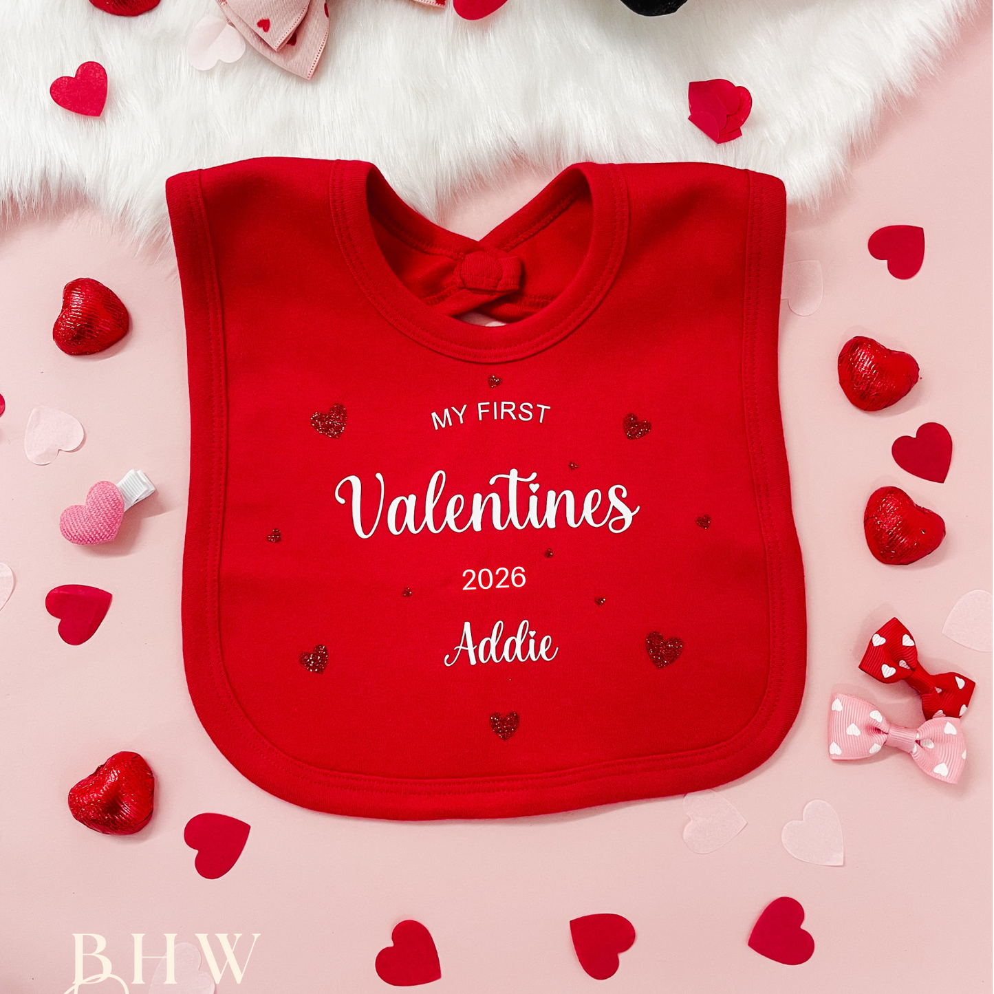 Red bib with 'My First Valentine's 2026' text, surrounded by heart decorations on a pink background.