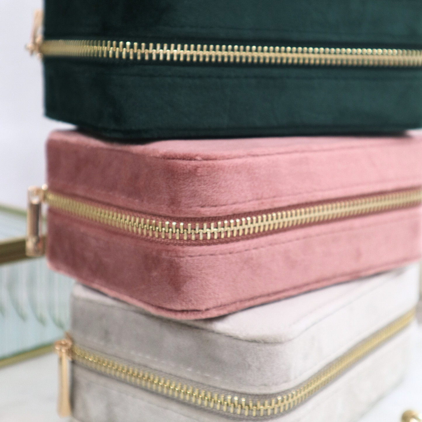 A stack of three velvet jewellery cases in rose quartz, green, and silver colors, with gold zippers.