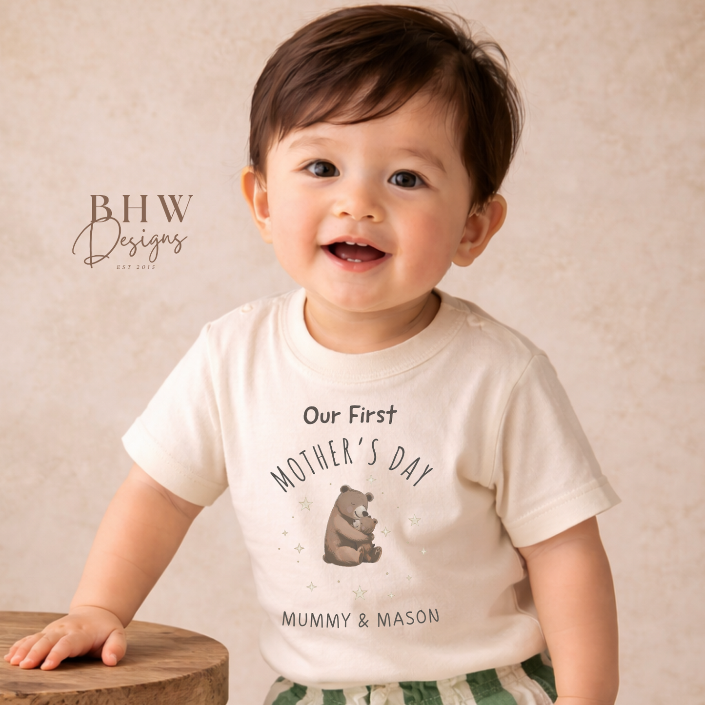Child wearing a 'Our First Mother's Day' shirt with a bear graphic, standing on a wooden stool.