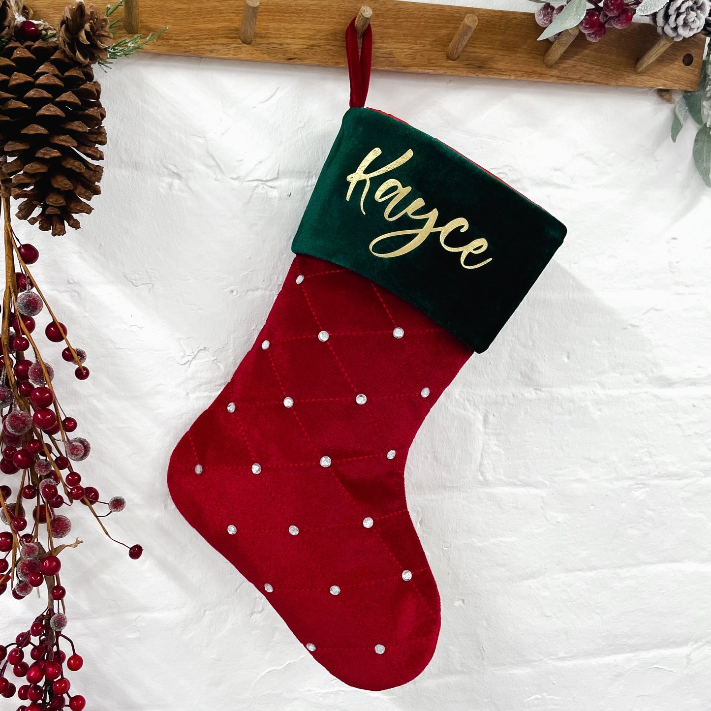 Red Christmas stocking with green top and 'Kayce' personalised surrounded by pine cones and berries on a white background.