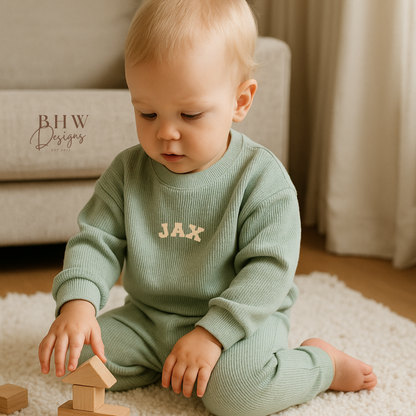 Baby playing with wooden blocks on a rug, wearing a sage green outfit with 'JAX' printed on it.