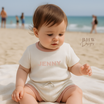 Baby sitting on a beach with in a white ribbed outfit with baby pink trim and  'JENNY' printed on the front in baby pink