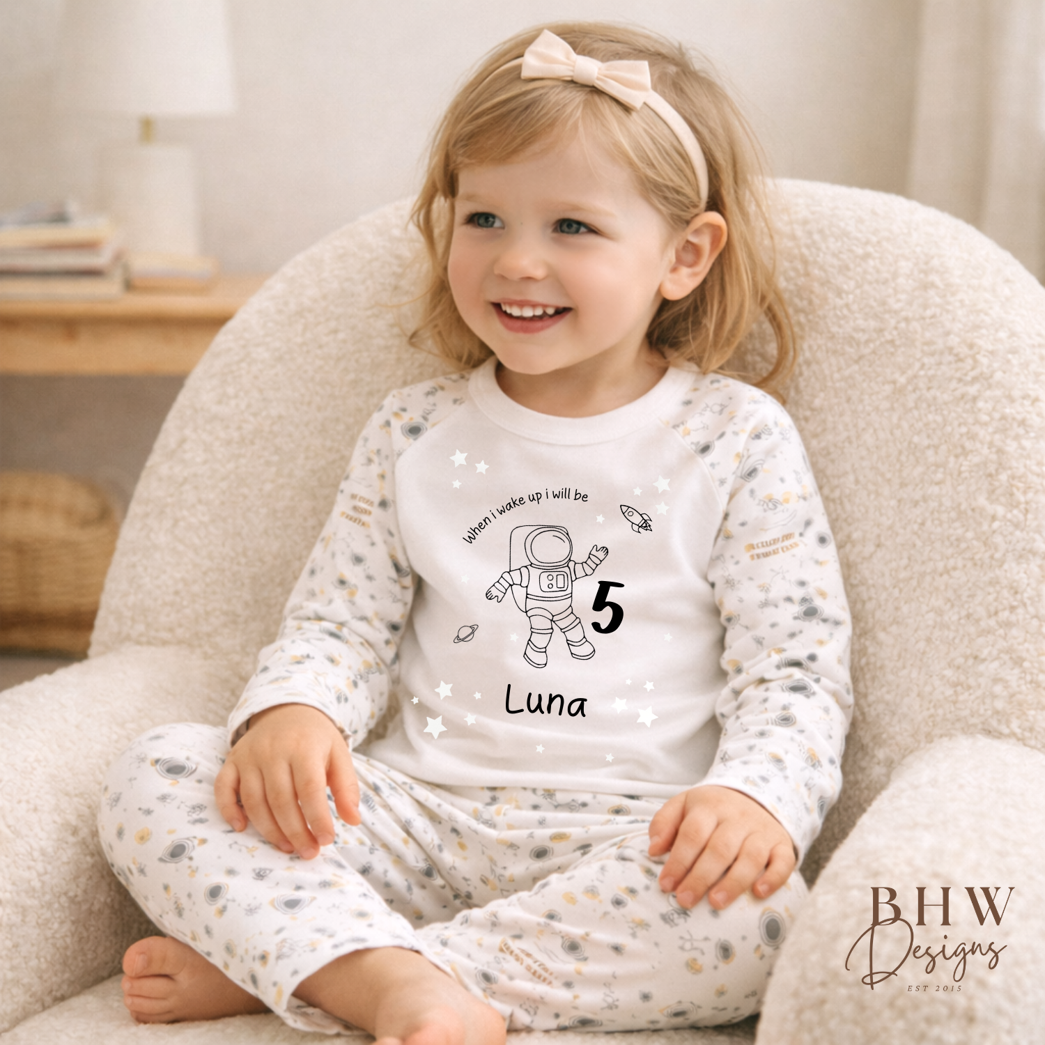 Child wearing space theme pyjamas with an astronaut design and text, sitting on a beige chair.