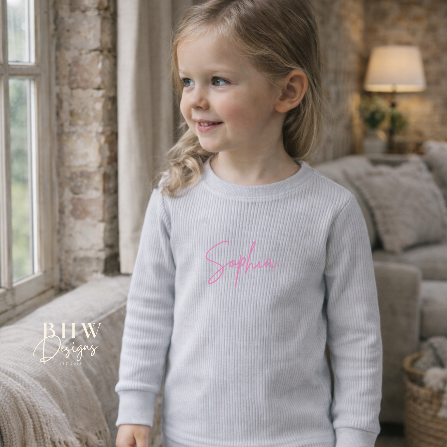 Child wearing a light grey ribbed loungewear set with 'Sophia' printed on the front standing in a cozy living room.