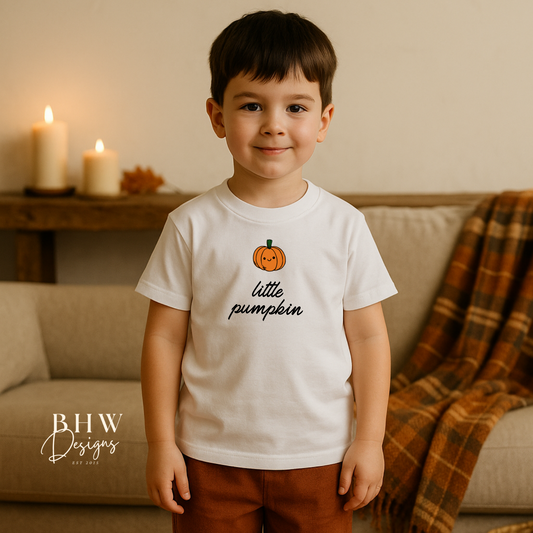 Child wearing a 'little pumpkin' shirt in a cozy living room setting with pumpkins and candles.