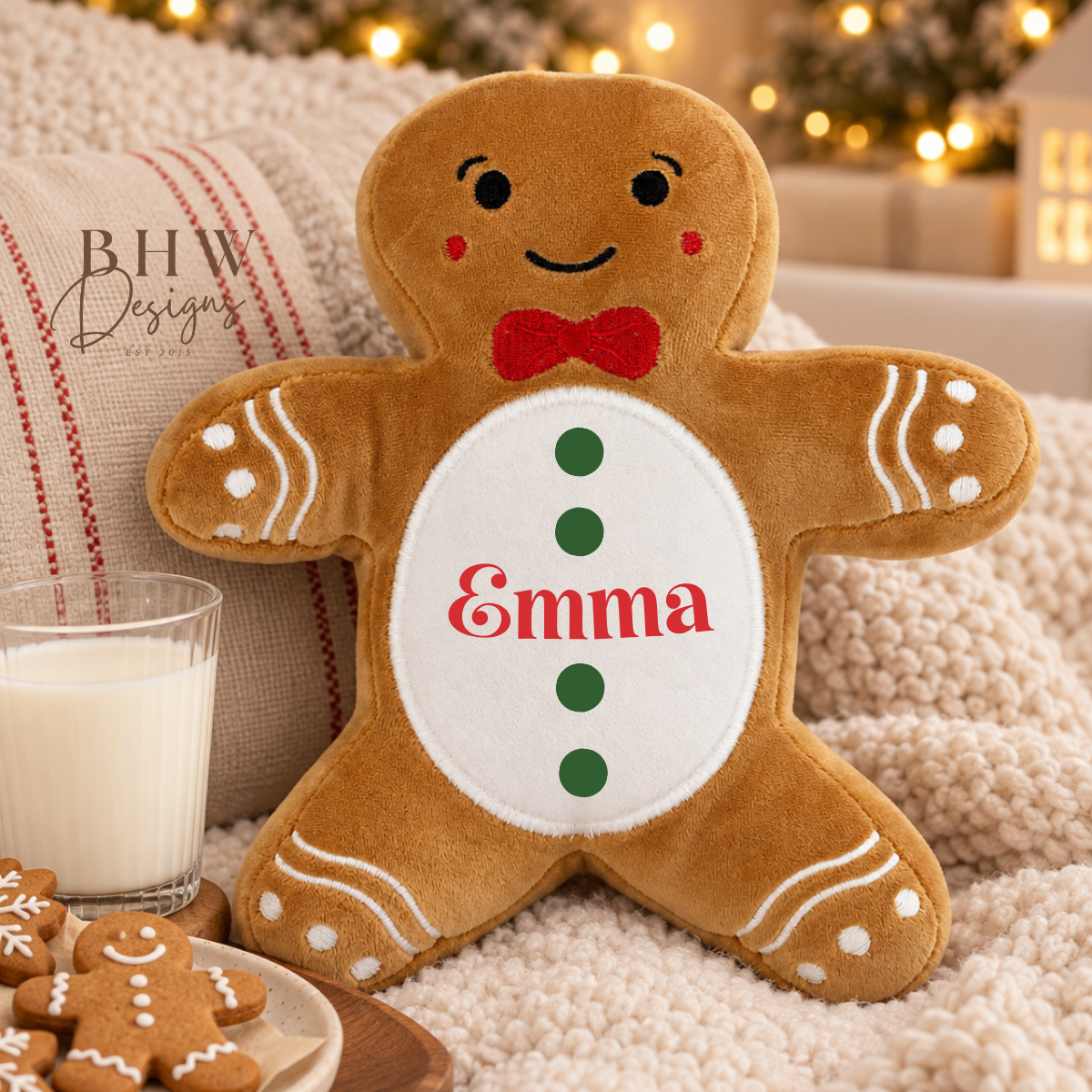 A plush gingerbread person pillow personalized with the name 'Emma' in red, styled on a cozy blanket next to a glass of milk and cookies. The pillow features a brown body with white icing details, a red bow tie, and green buttons.