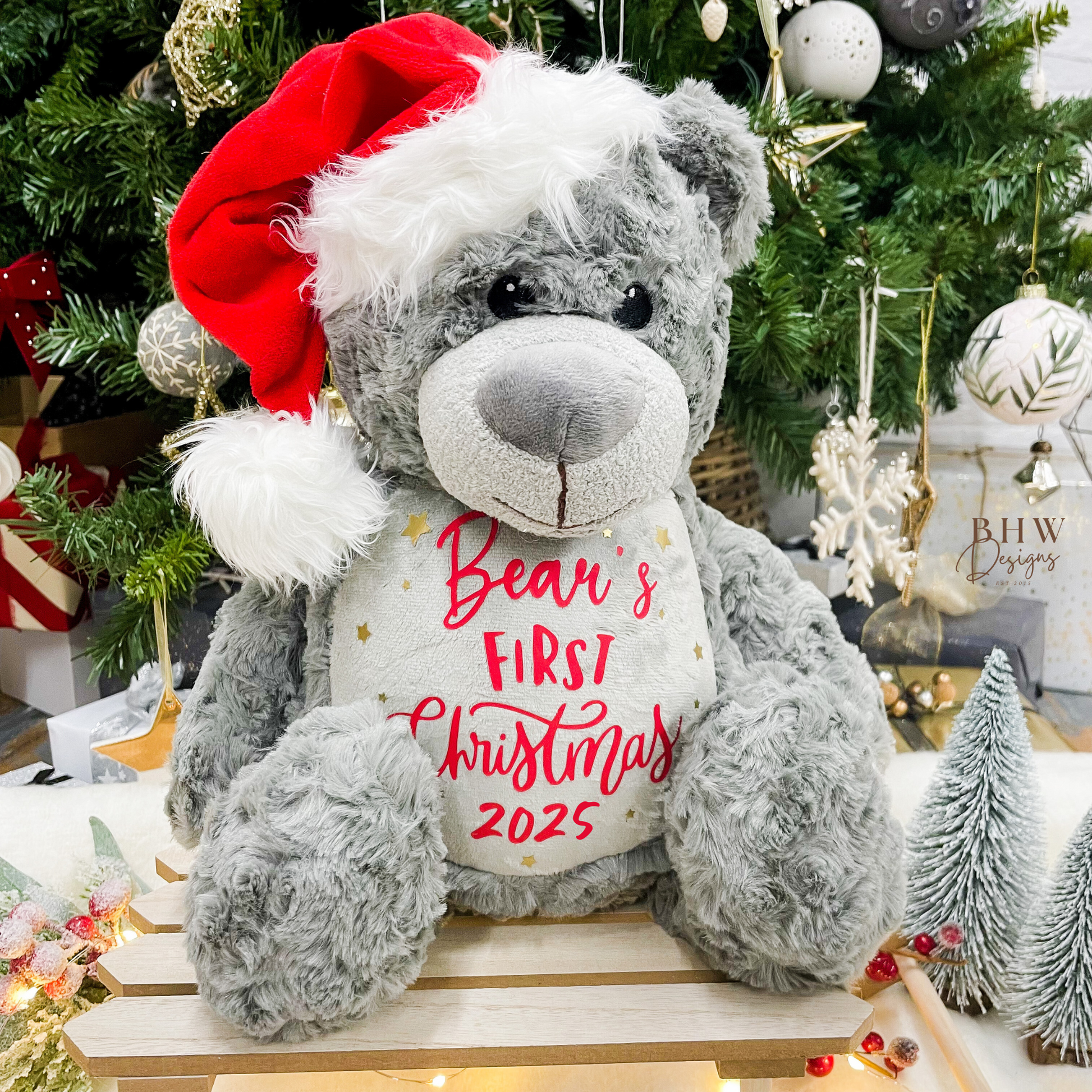 Grey teddy bear with a red Santa hat and 'Bear's First Christmas 2025' text in a festive setting.