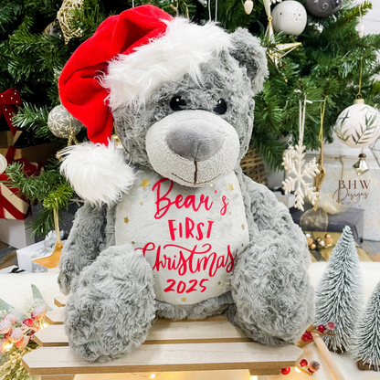 Grey teddy bear with a red Santa hat and 'Bear's First Christmas 2025' text in a festive setting.
