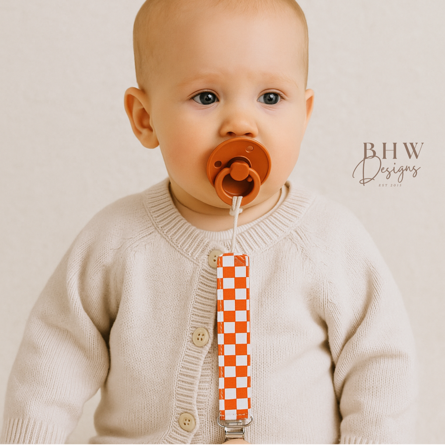 Baby wearing an orange pacifier with a checkered strap against a plain background