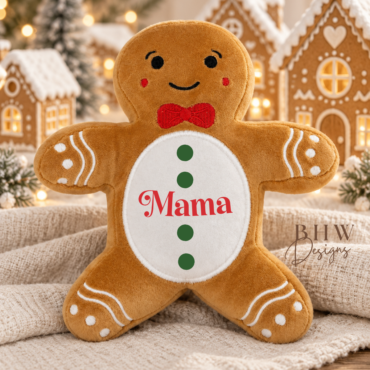A plush gingerbread person pillow with the name 'Mama' embroidered on its belly, styled on a beige knit blanket with a cozy Christmas background of gingerbread houses and warm lights.
