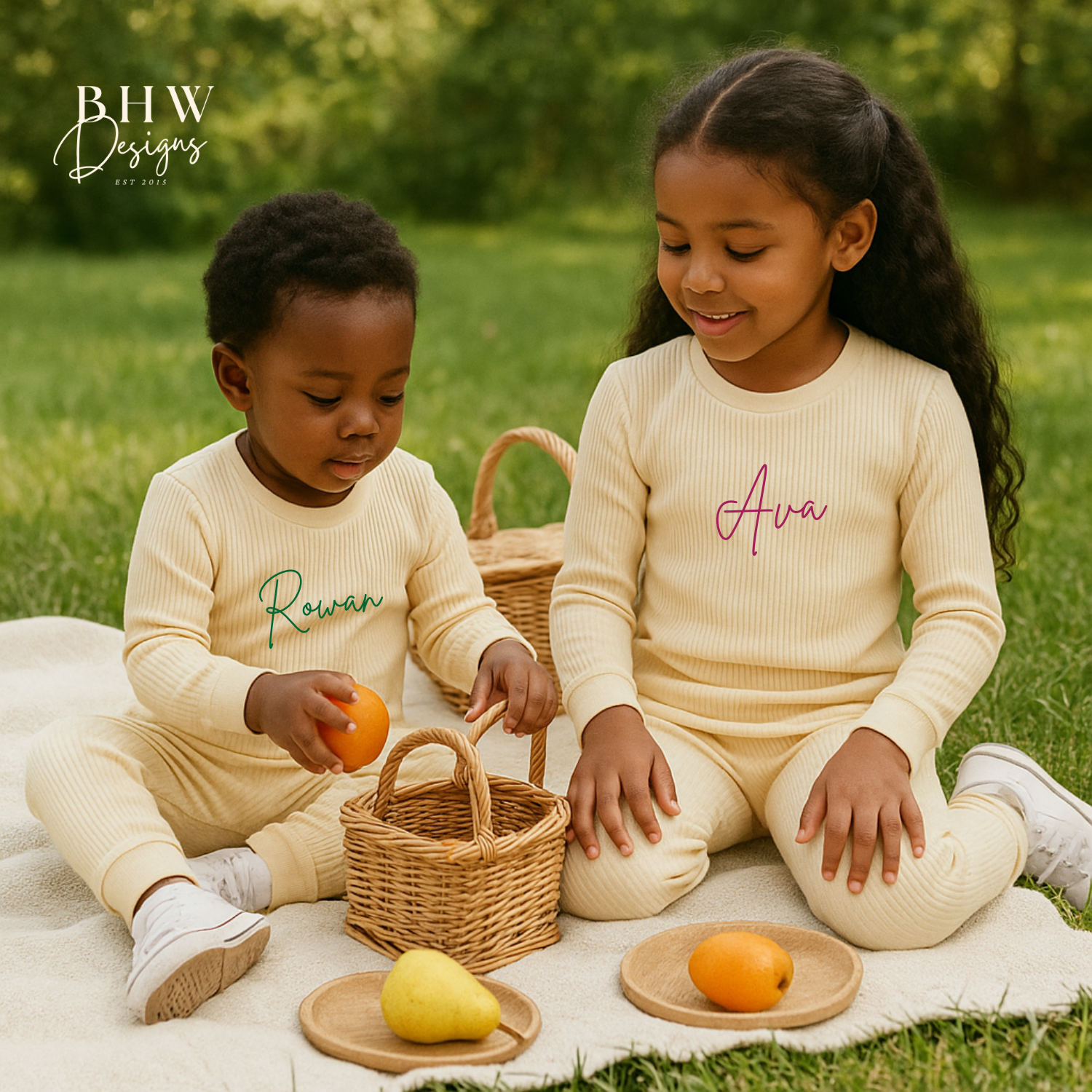 Two children sitting on a blanket outdoors with baskets and fruits, wearing matching outfits cream loungewear with names printed.