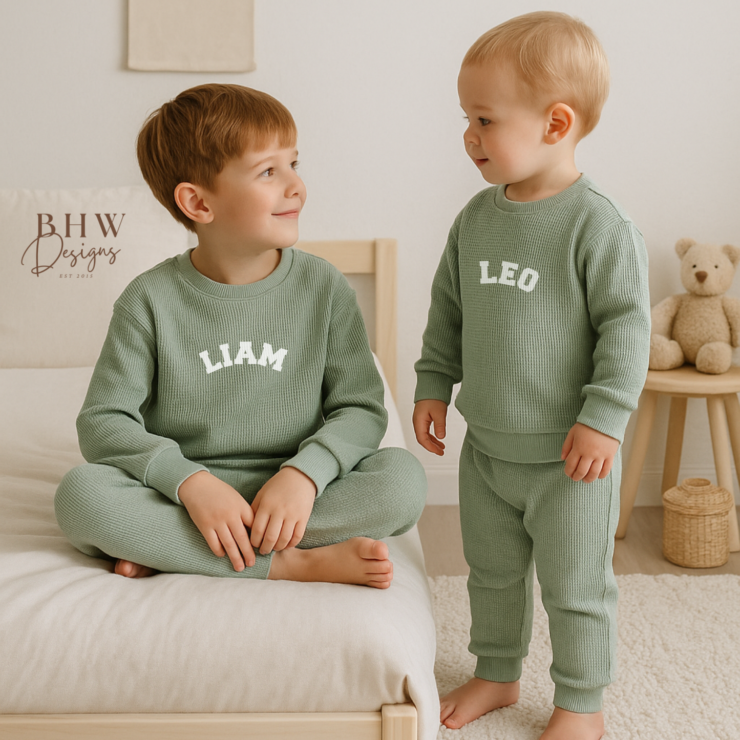 Two children wearing matching sage green outfits with names printed on them, sitting on a bed.