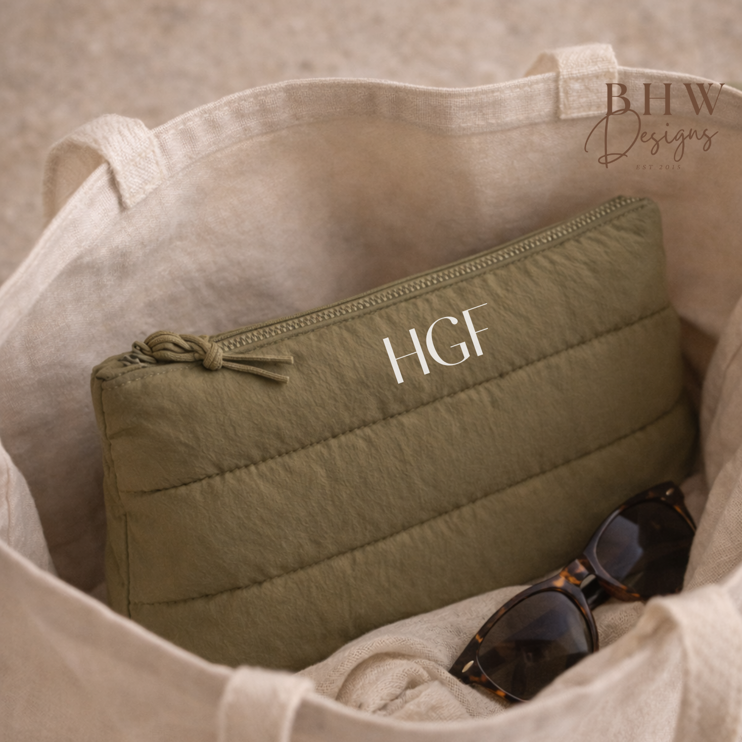 Green quilted pouch with 'HGF' monogram inside a beige bag, with sunglasses on a neutral background.