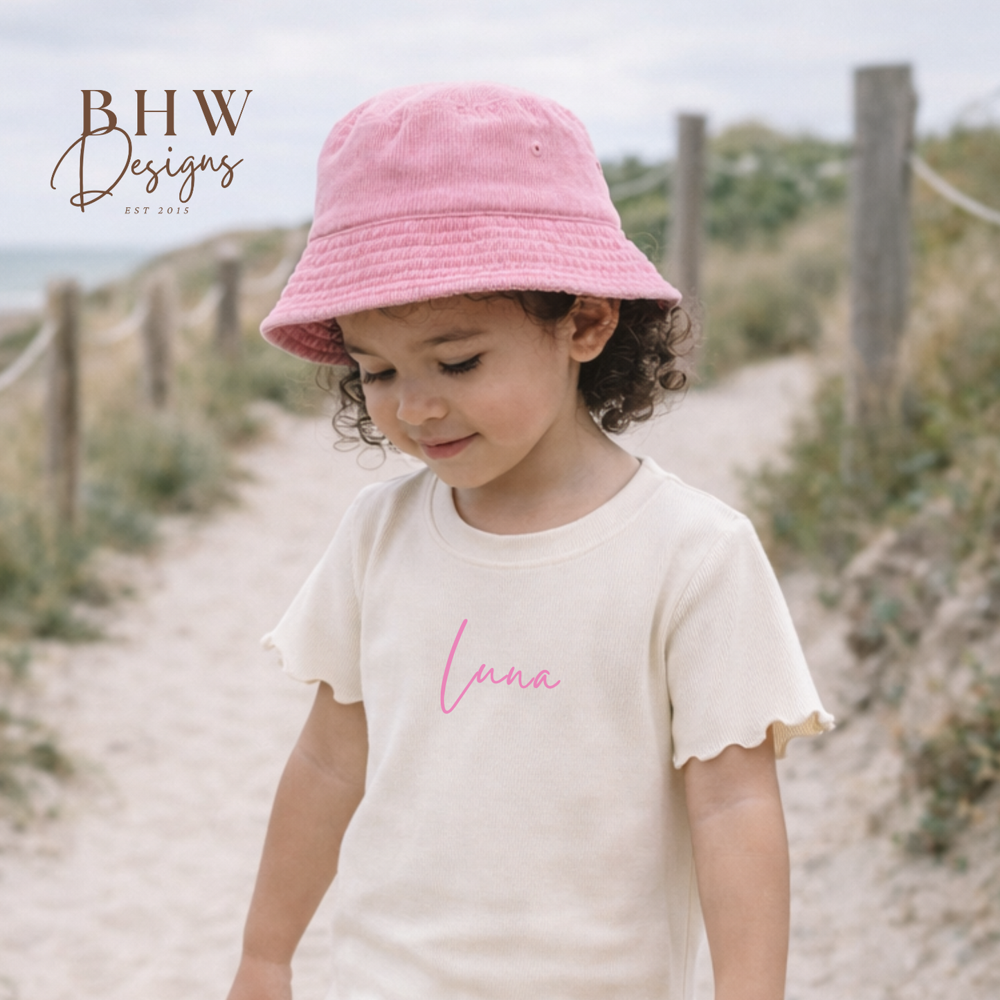 Child wearing a pink bucket hat and cream outfit with 'Lana' printed on it, standing on a sandy path.