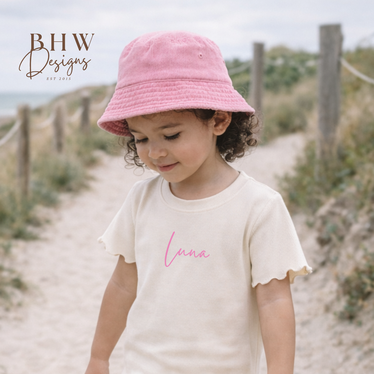 Child wearing a pink bucket hat and cream outfit with 'Lana' printed on it, standing on a sandy path.