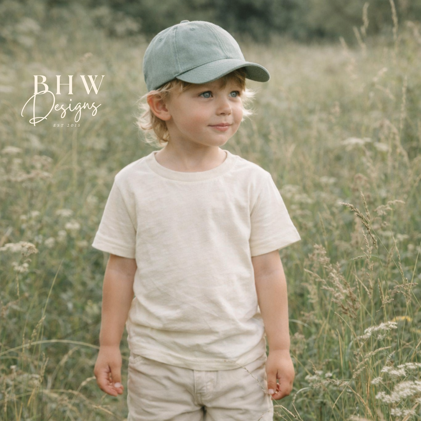 Young boy in a field wearing a cap, t-shirt, shorts, and rain boots.