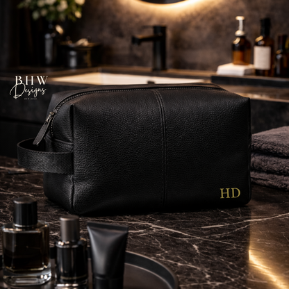 Black toiletry bag with 'HD' initials on a marble bathroom counter with grooming products.