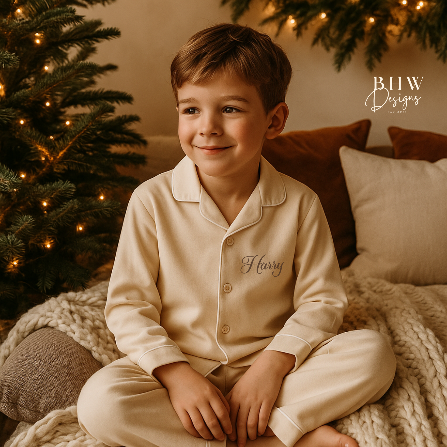 Boy in beige cotton pyjamas Personalised with Harry in Brown sitting in front of a decorated Christmas tree.