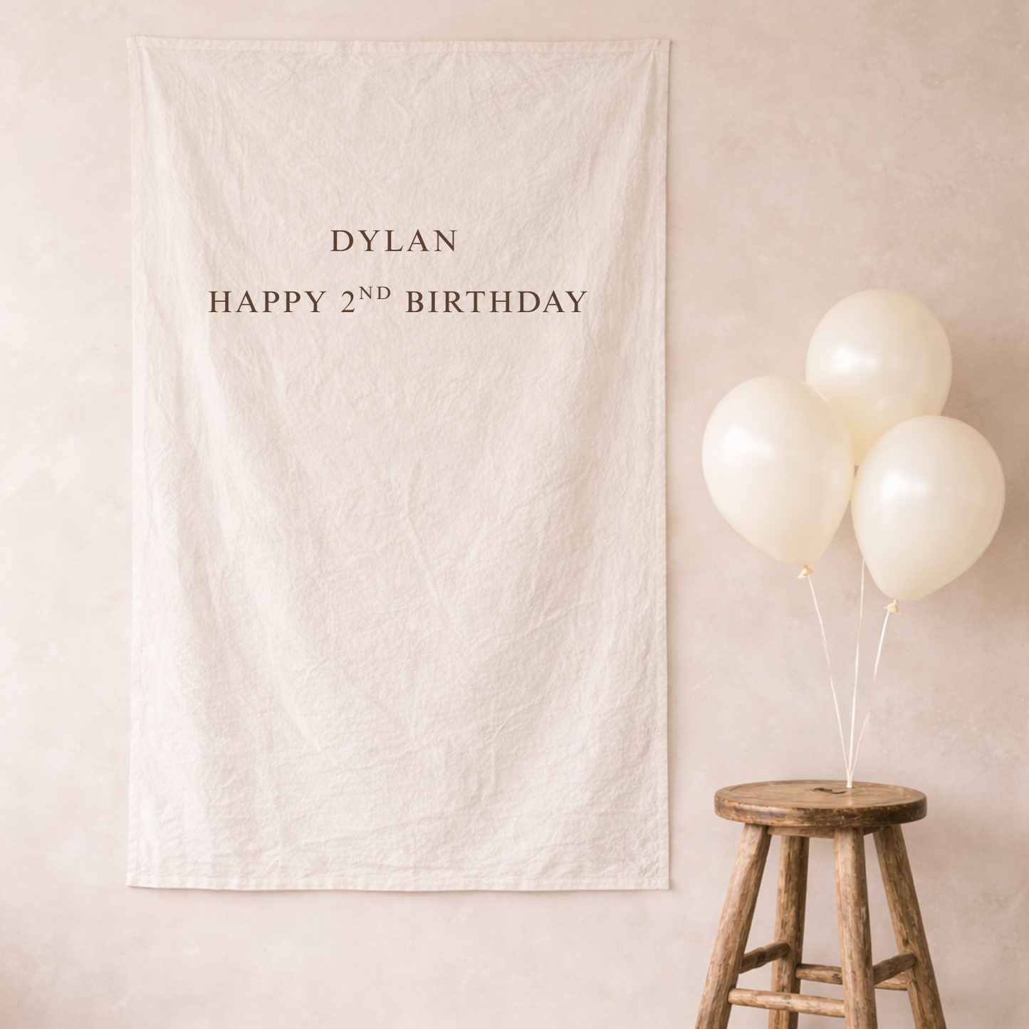 Beige banner with 'Dylan Happy 2nd Birthday' text, white balloons on a wooden stool against a beige wall.